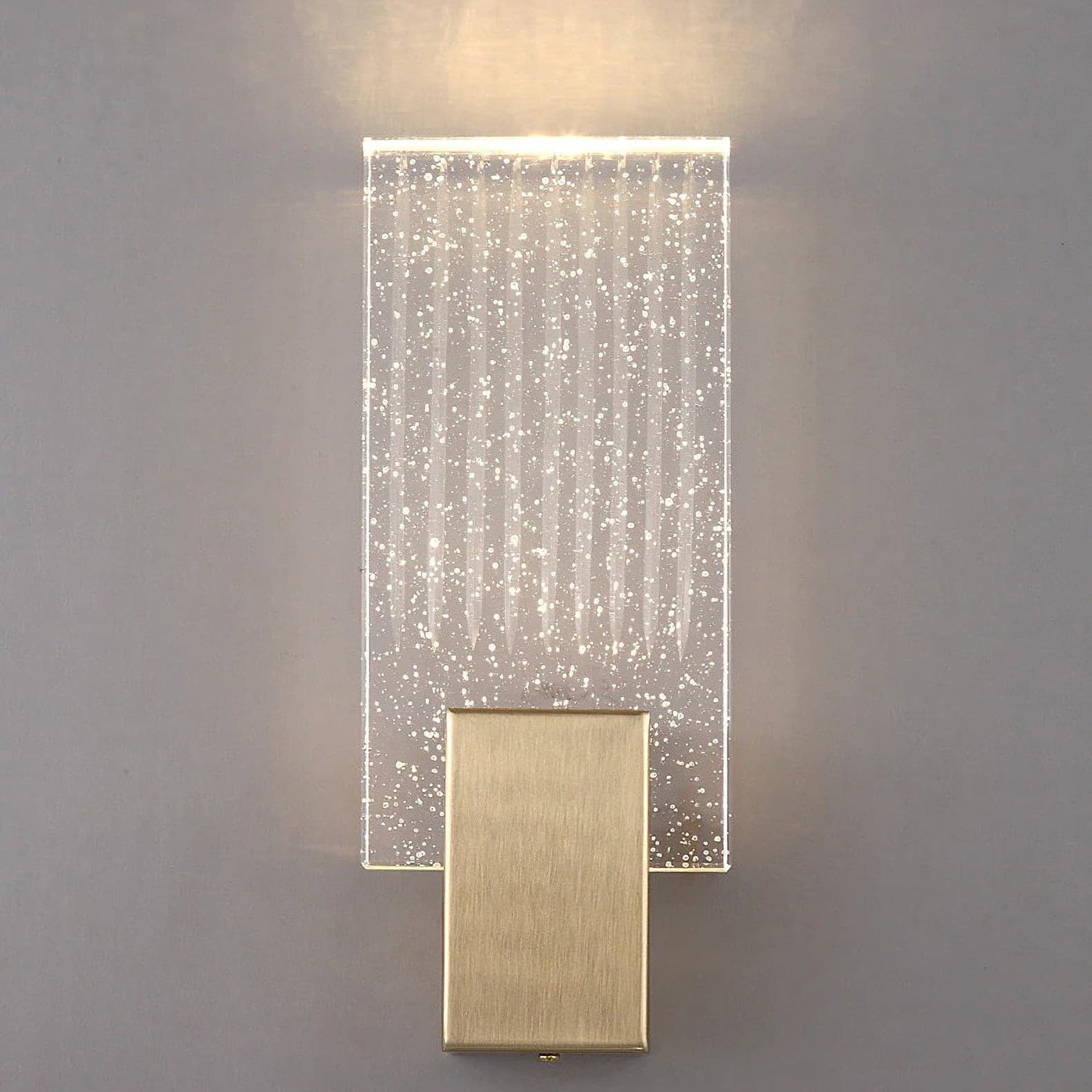Wall light with a ribbed clear glass panel and a brushed brass mount lit, shown close on a plain wall.