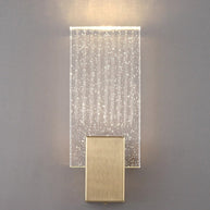 Wall light with a ribbed clear glass panel and a brushed brass mount lit, shown close on a plain wall.
