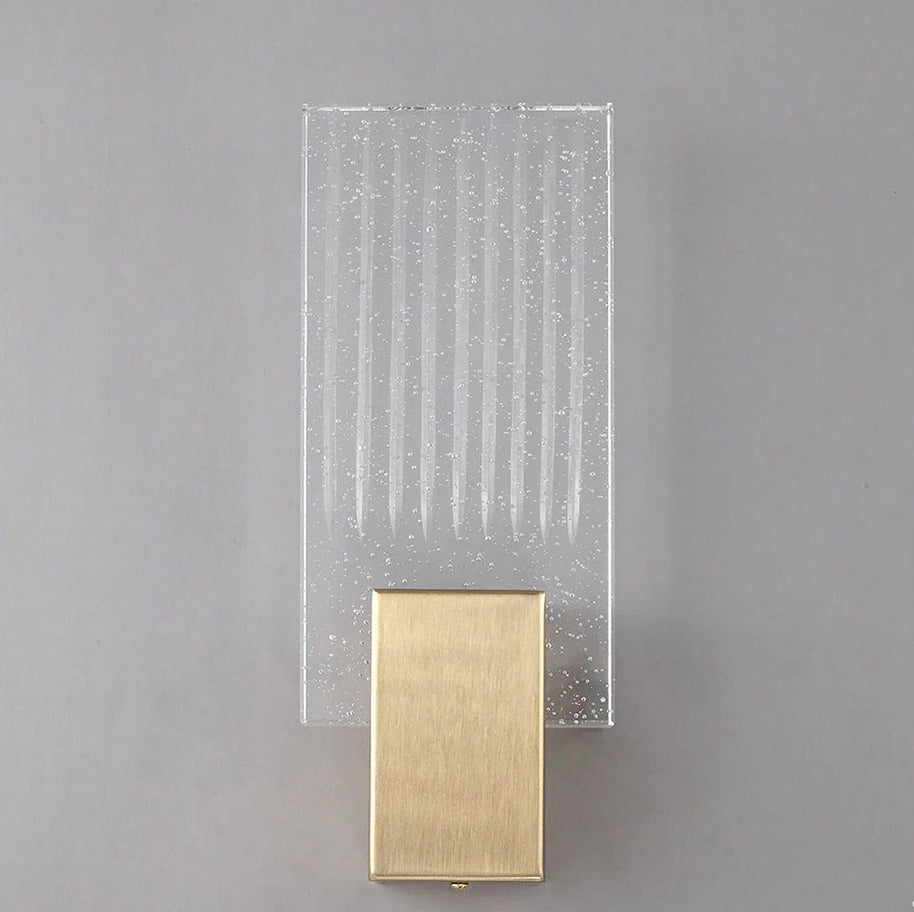 Wall light with a ribbed clear glass panel and a brushed brass mount unlit, shown close on a plain wall.