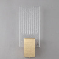 Wall light with a ribbed clear glass panel and a brushed brass mount unlit, shown close on a plain wall.