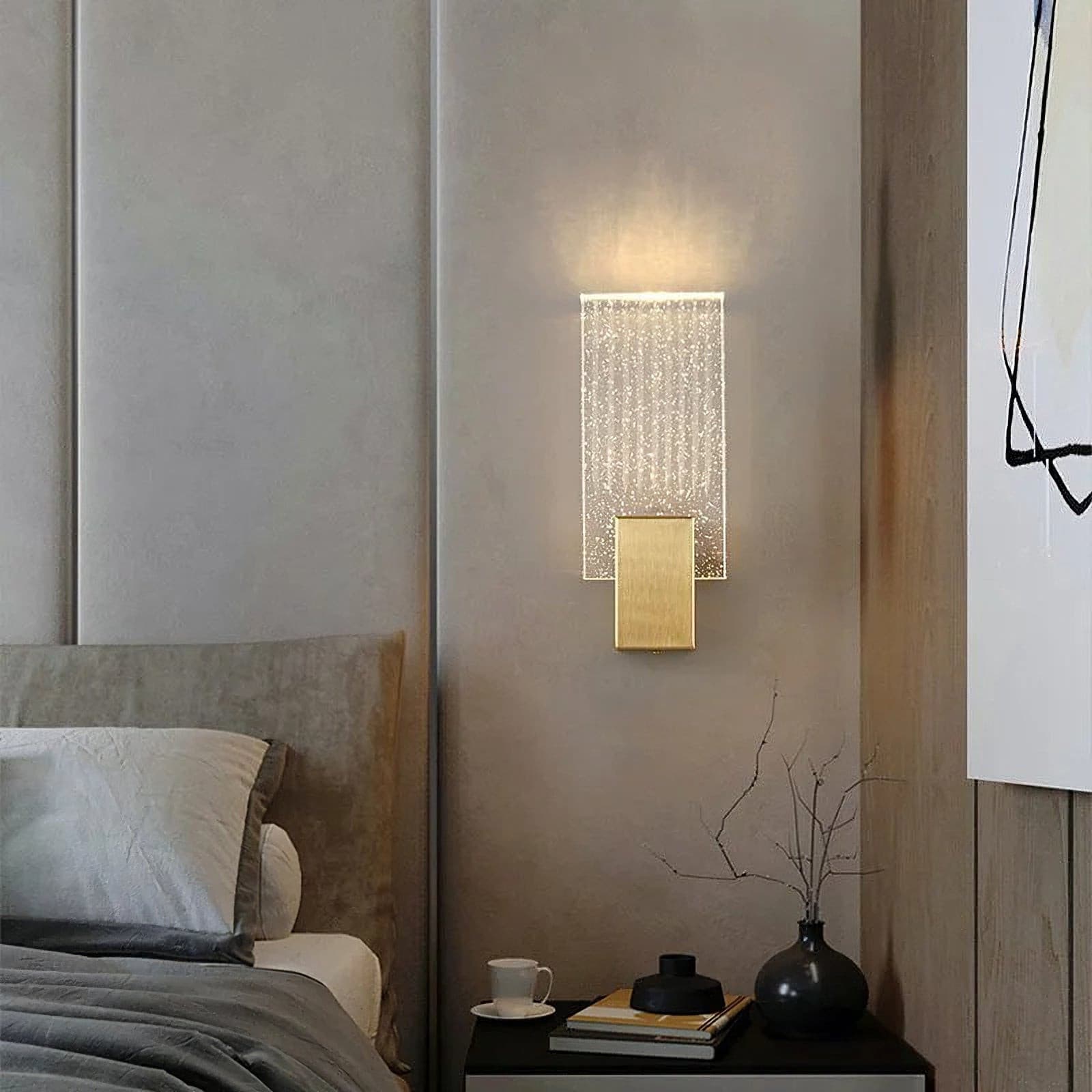 Wall light with a ribbed clear glass panel and a brushed brass mount lit on a bedroom wall beside a bed.