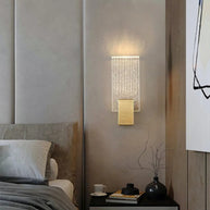 Wall light with a ribbed clear glass panel and a brushed brass mount lit on a bedroom wall beside a bed.