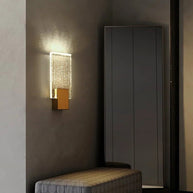 Wall light with a ribbed clear glass panel and a brushed brass mount lit on a wall beside a doorway and a bench.