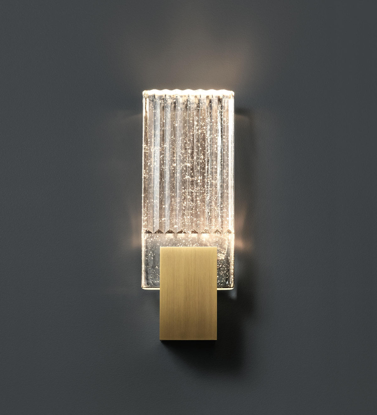 Wall light with a ribbed clear glass panel and a brushed brass mount lit against a dark grey wall.