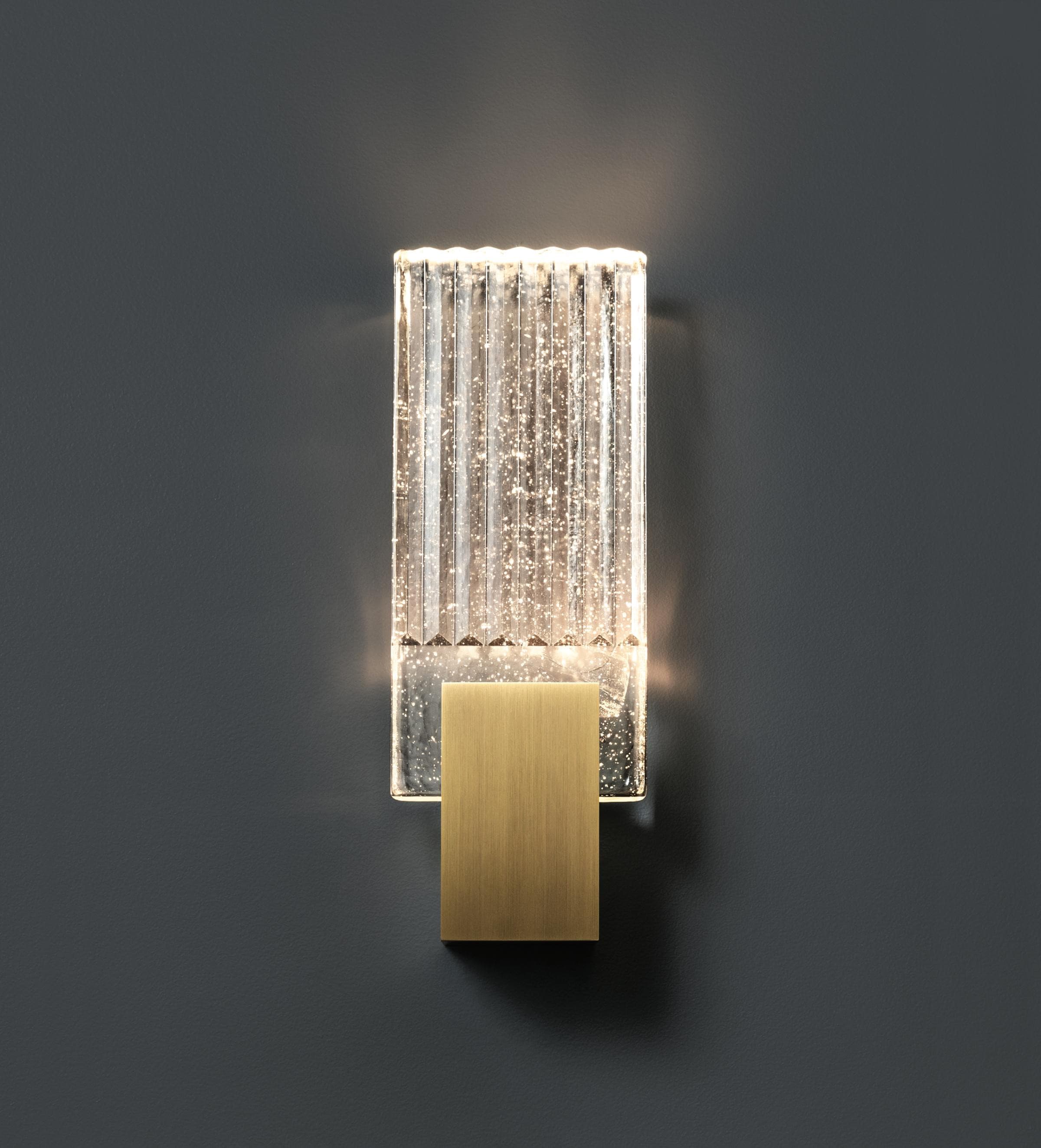 Wall light with a ribbed clear glass panel and a brushed brass mount lit against a dark grey wall.