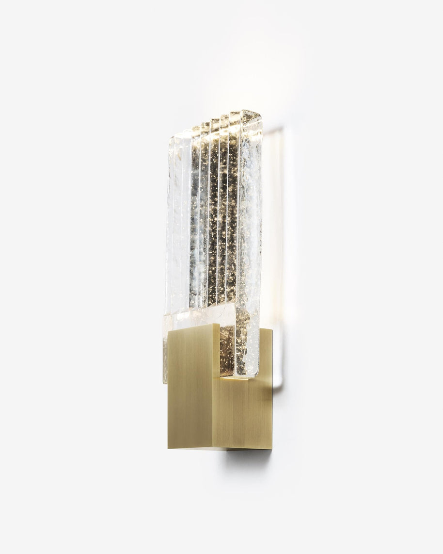 Wall light with a ribbed clear glass panel and a brushed brass mount seen at an angle, lit, on a white background.