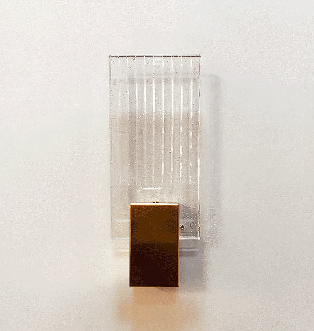Wall light with a ribbed clear glass panel and a brushed brass mount, seen at an angle on a pale wall.