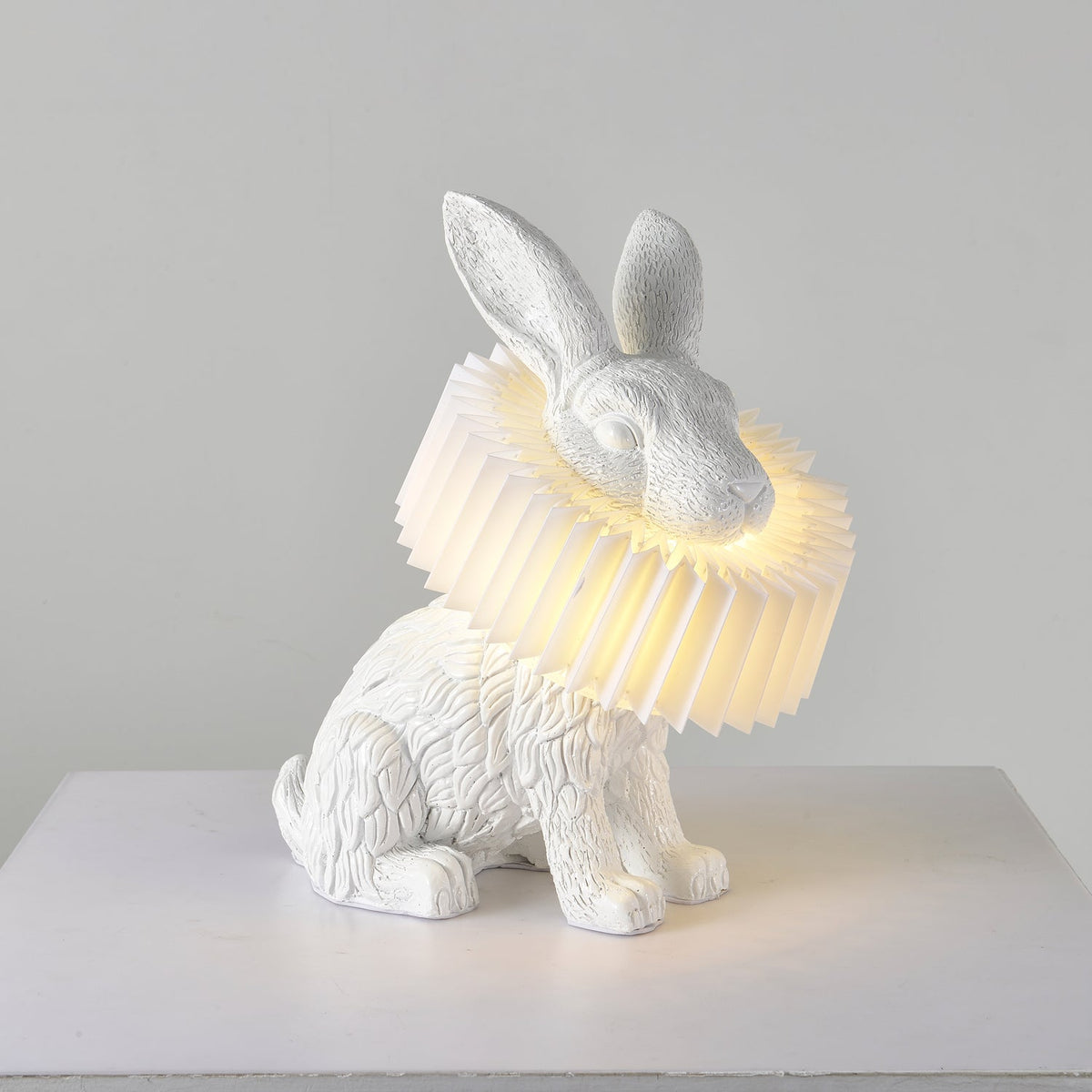 Rabellin Table Lamp - seated white leaf-textured rabbit with pleated ruff shade glowing warm on a white plinth