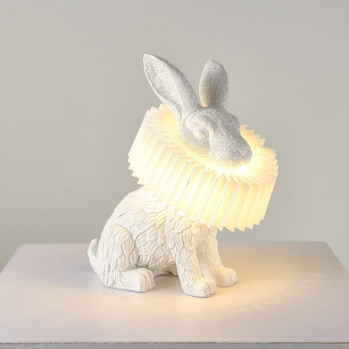 Rabellin Table Lamp - seated white leaf-textured rabbit with pleated ruff shade glowing warm on a white plinth