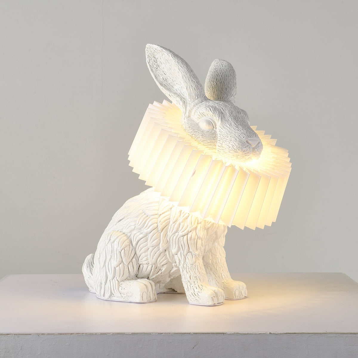 Rabellin Table Lamp - seated white leaf-textured rabbit with pleated ruff shade glowing warm on a white plinth