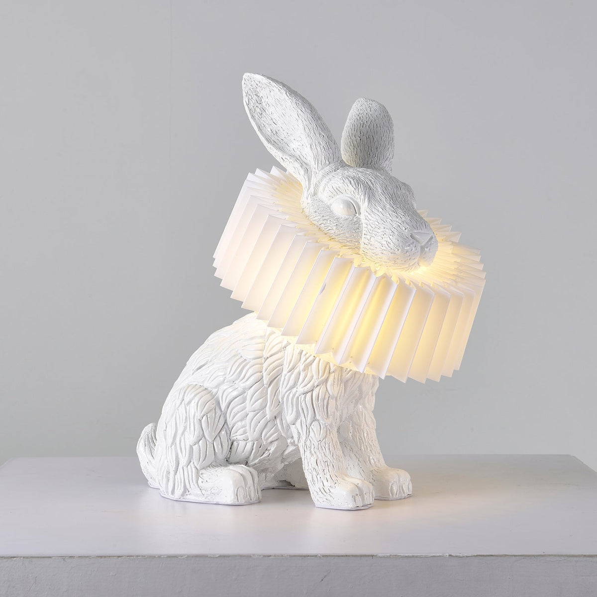 Rabellin Table Lamp - seated white leaf-textured rabbit with pleated ruff shade glowing warm on a white plinth