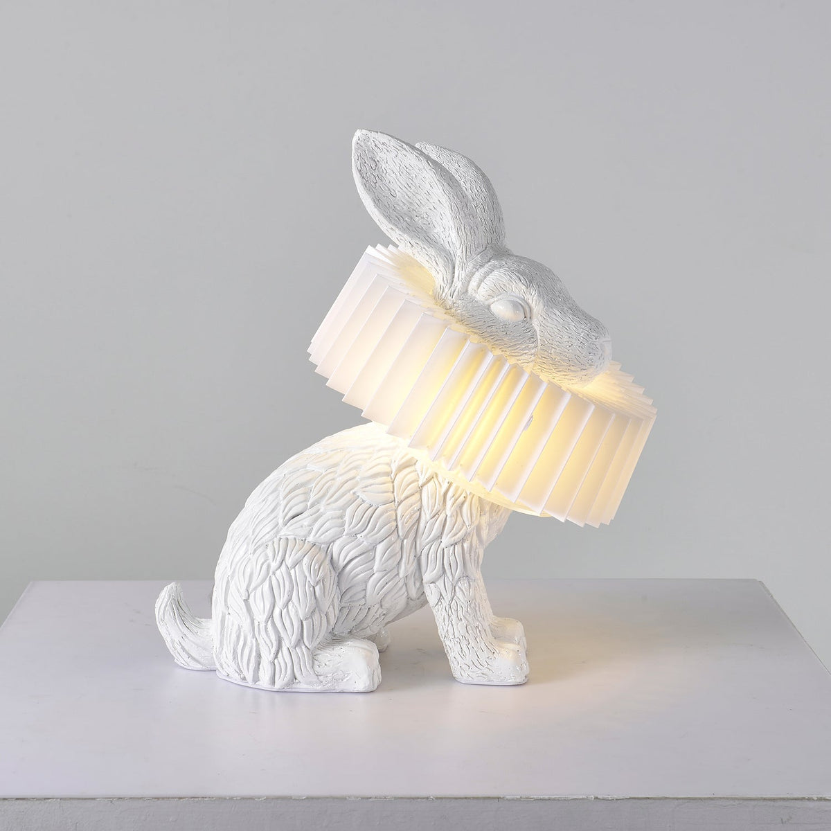 Rabellin Table Lamp - seated white leaf-textured rabbit with pleated ruff shade glowing warm on a white plinth