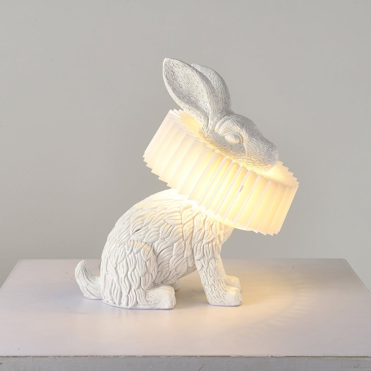 Rabellin Table Lamp - seated white leaf-textured rabbit with pleated ruff shade glowing warm on a white plinth