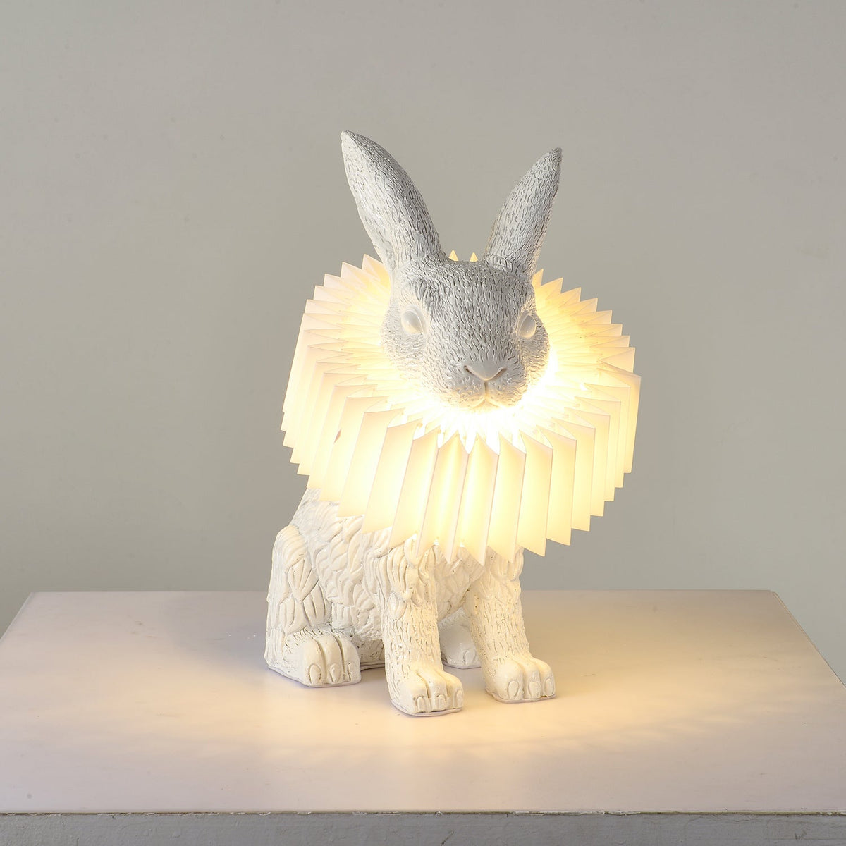 Rabellin Table Lamp - seated white leaf-textured rabbit with pleated ruff shade glowing warm on a white plinth