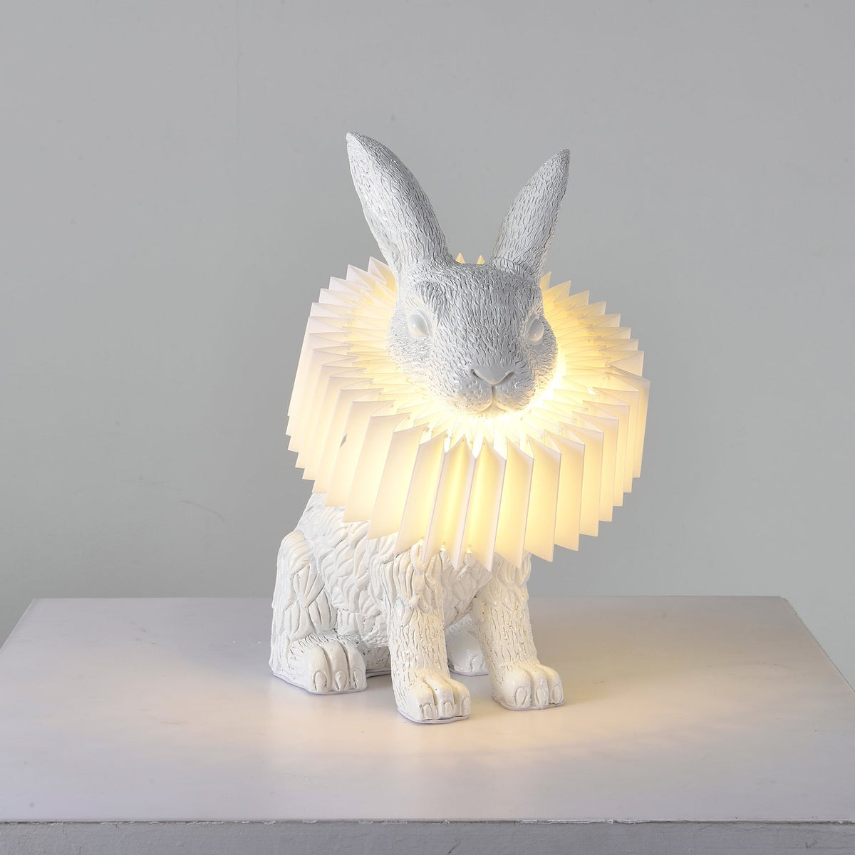Rabellin Table Lamp - seated white leaf-textured rabbit with pleated ruff shade glowing warm on a white plinth