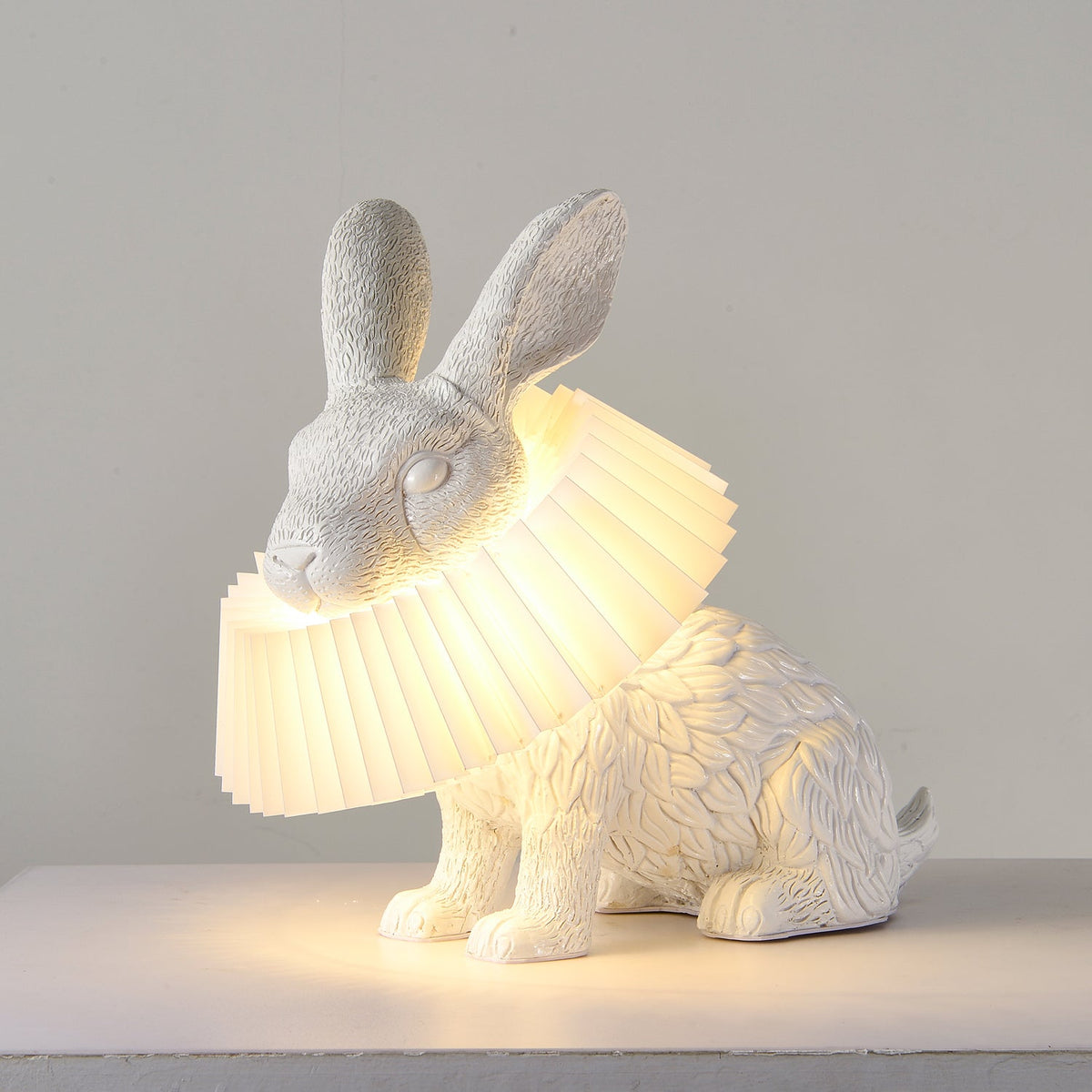 Rabellin Table Lamp - seated white leaf-textured rabbit with pleated ruff shade glowing warm on a white plinth