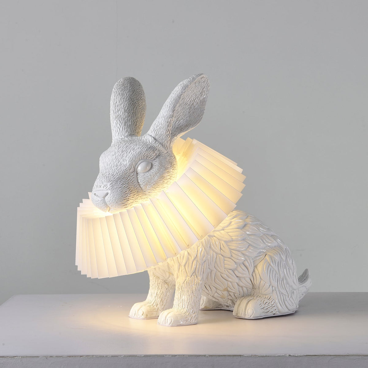 Rabellin Table Lamp - seated white leaf-textured rabbit with pleated ruff shade glowing warm on a white plinth