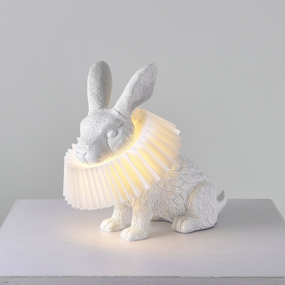 Rabellin Table Lamp - seated white leaf-textured rabbit with pleated ruff shade glowing warm on a white plinth
