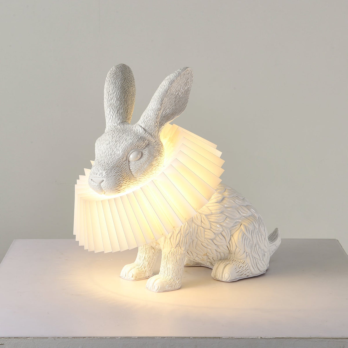 Rabellin Table Lamp - seated white leaf-textured rabbit with pleated ruff shade glowing warm on a white plinth
