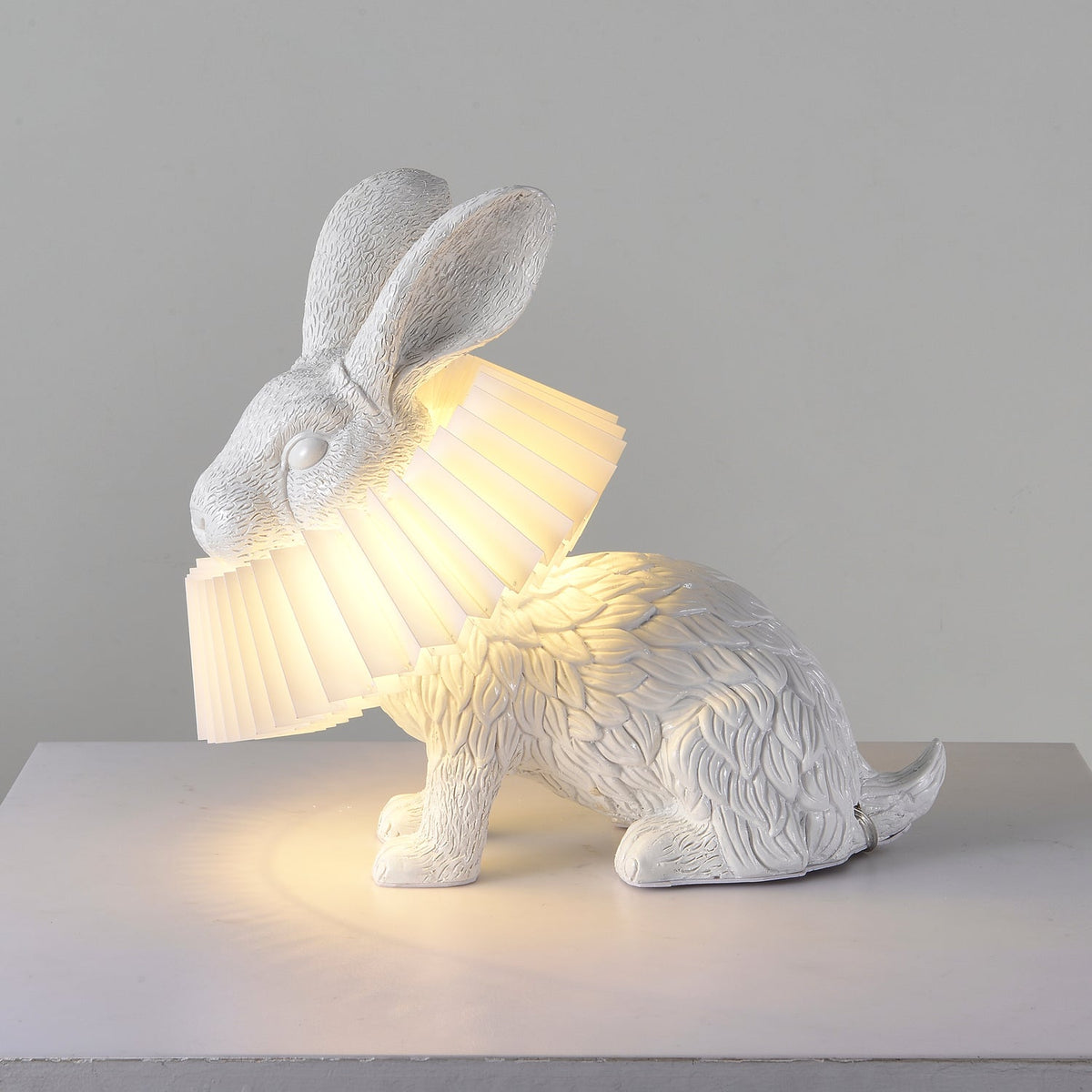 Rabellin Table Lamp - seated white leaf-textured rabbit with pleated ruff shade glowing warm on a white plinth