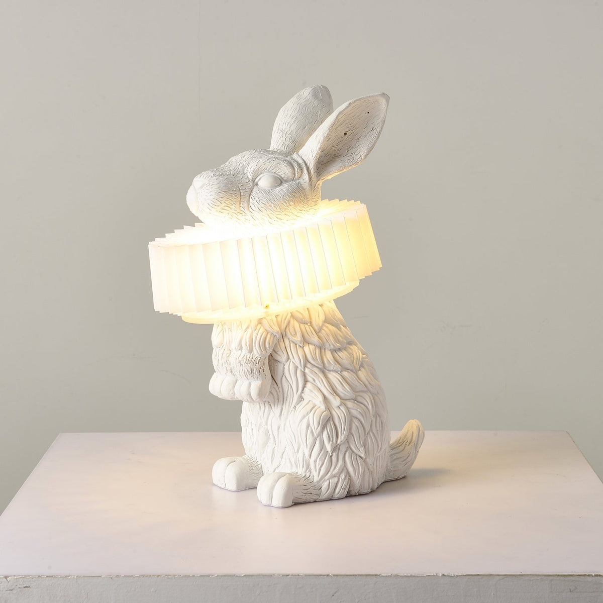 Rabellin Table Lamp - seated white leaf-textured rabbit with pleated ruff shade glowing warm on a white plinth