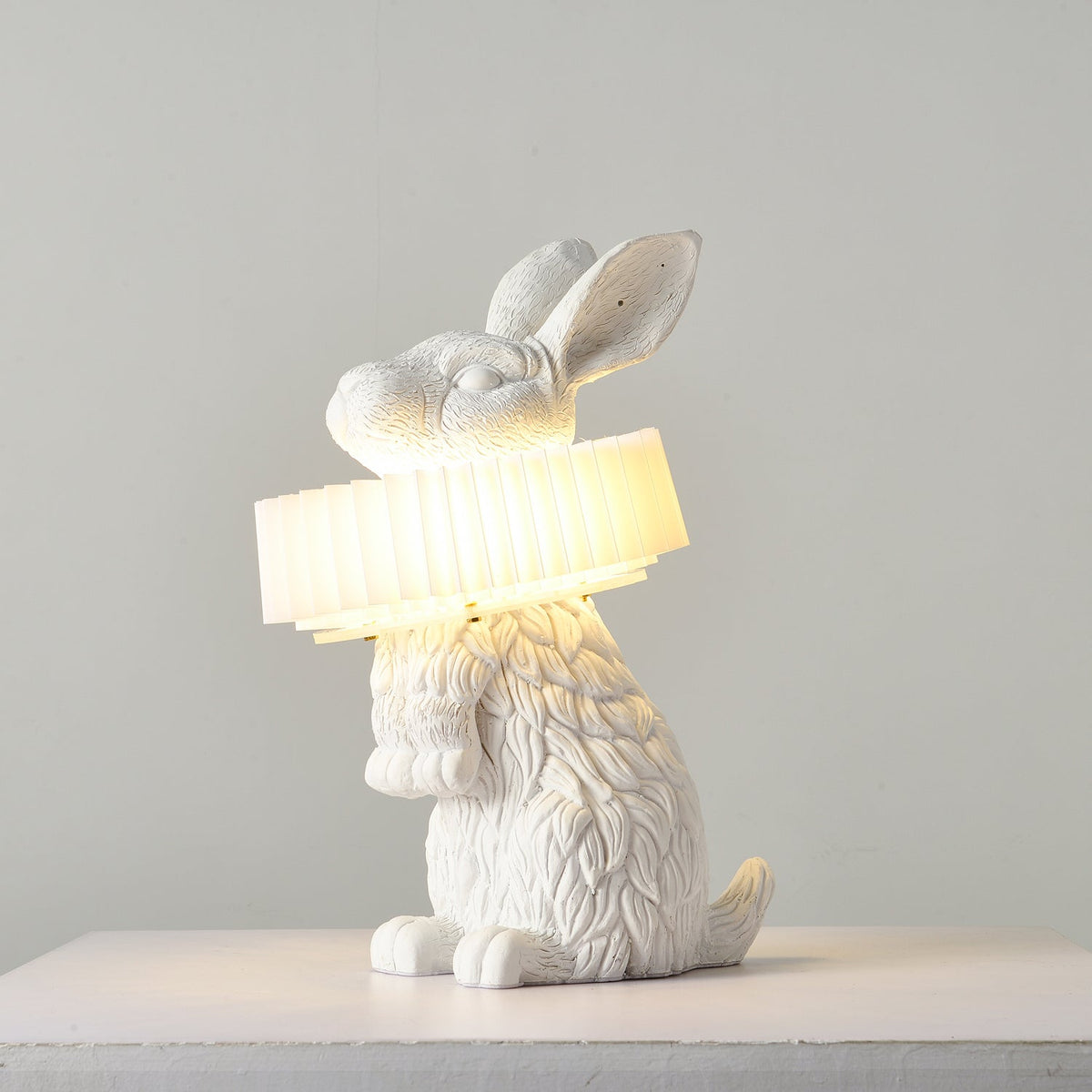 Rabellin Table Lamp - seated white leaf-textured rabbit with pleated ruff shade glowing warm on a white plinth