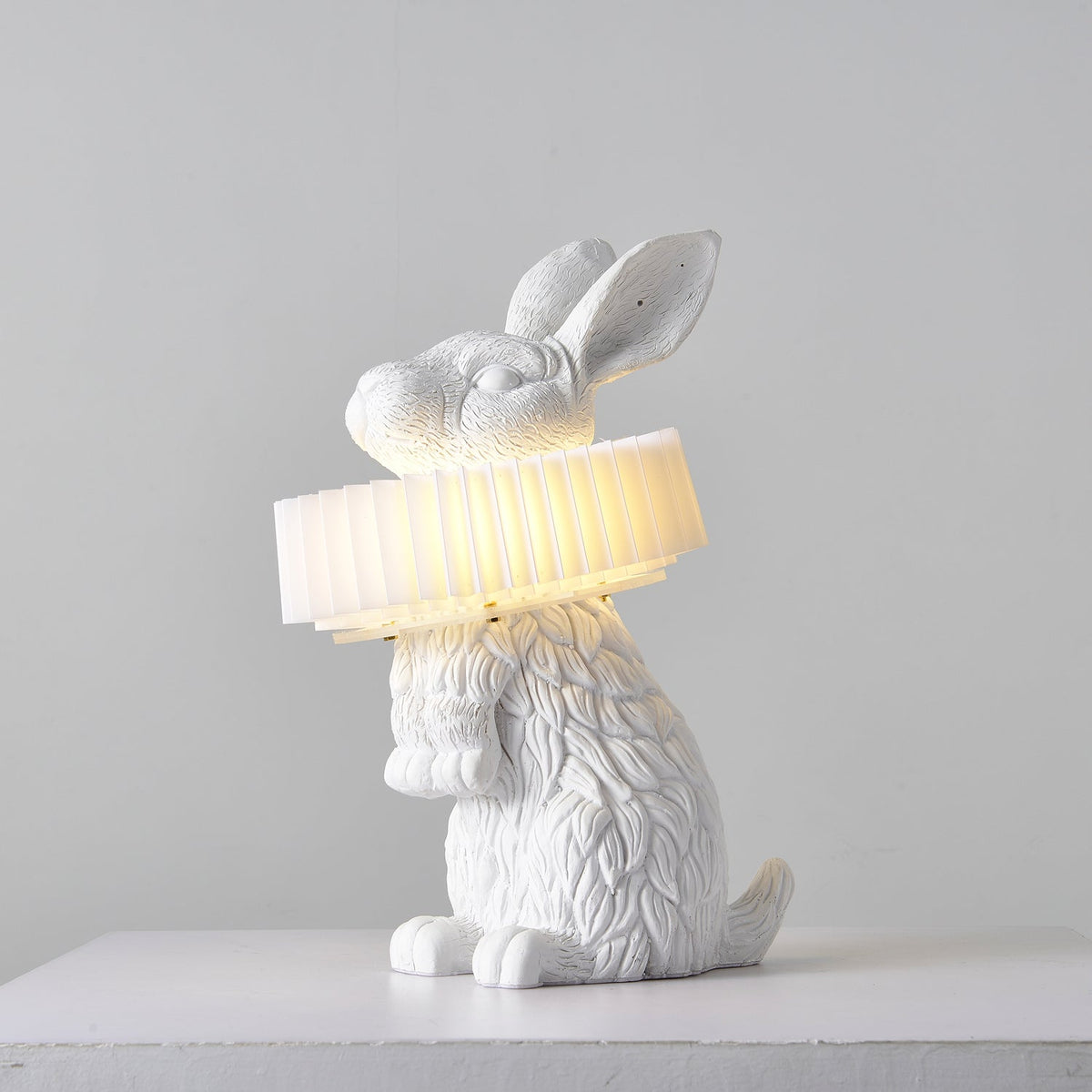Rabellin Table Lamp - seated white leaf-textured rabbit with pleated ruff shade glowing warm on a white plinth