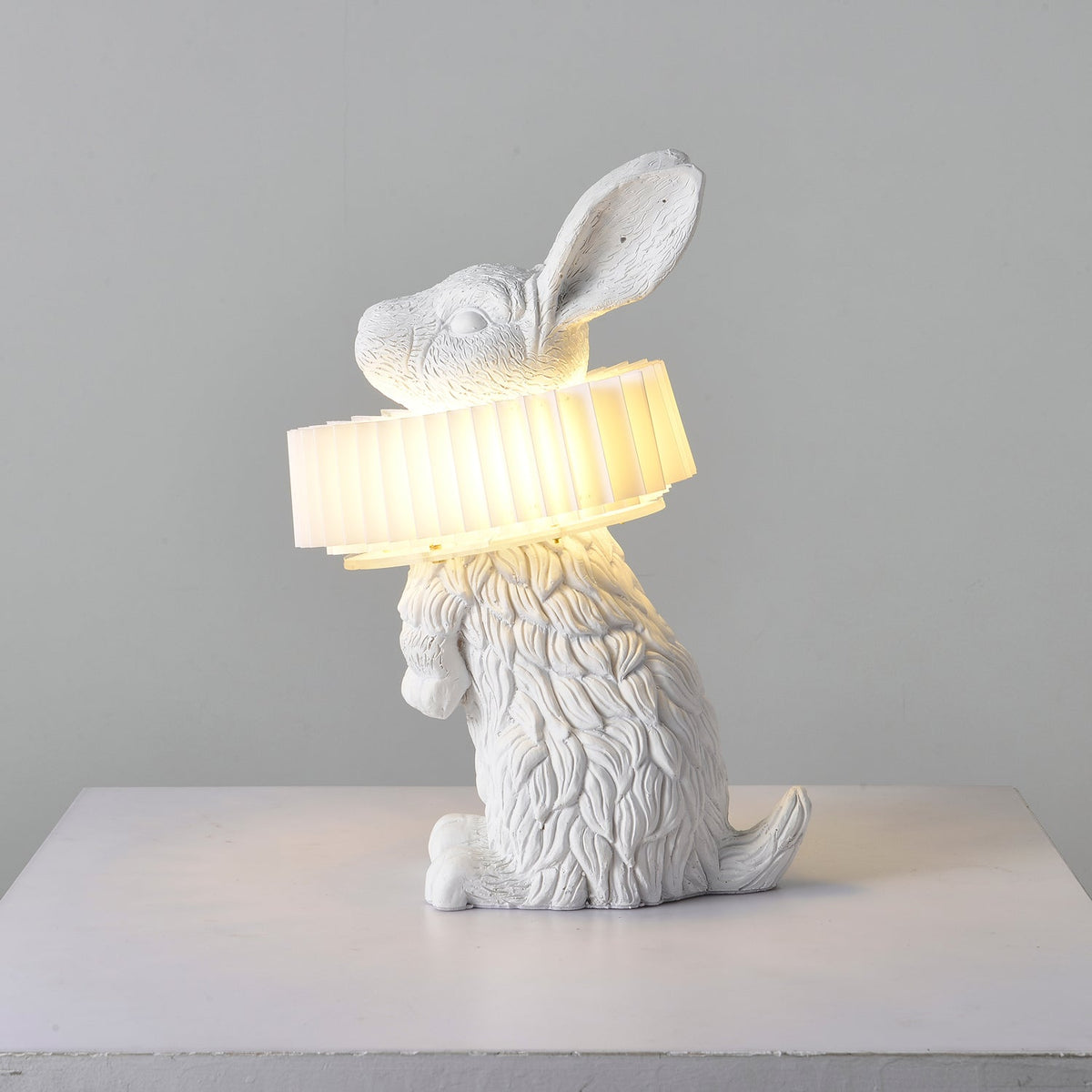 Rabellin Table Lamp - seated white leaf-textured rabbit with pleated ruff shade glowing warm on a white plinth