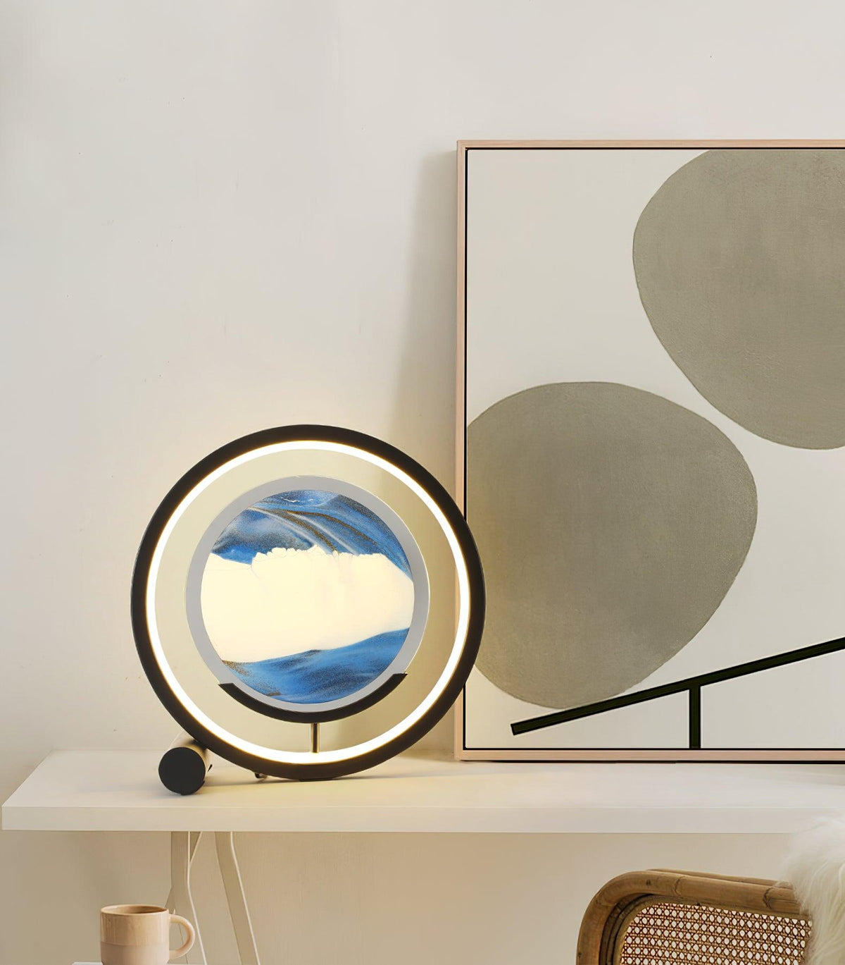 Sandrelle Table Lamp in black with lit LED ring and blue sand panel on a white shelf beside abstract art