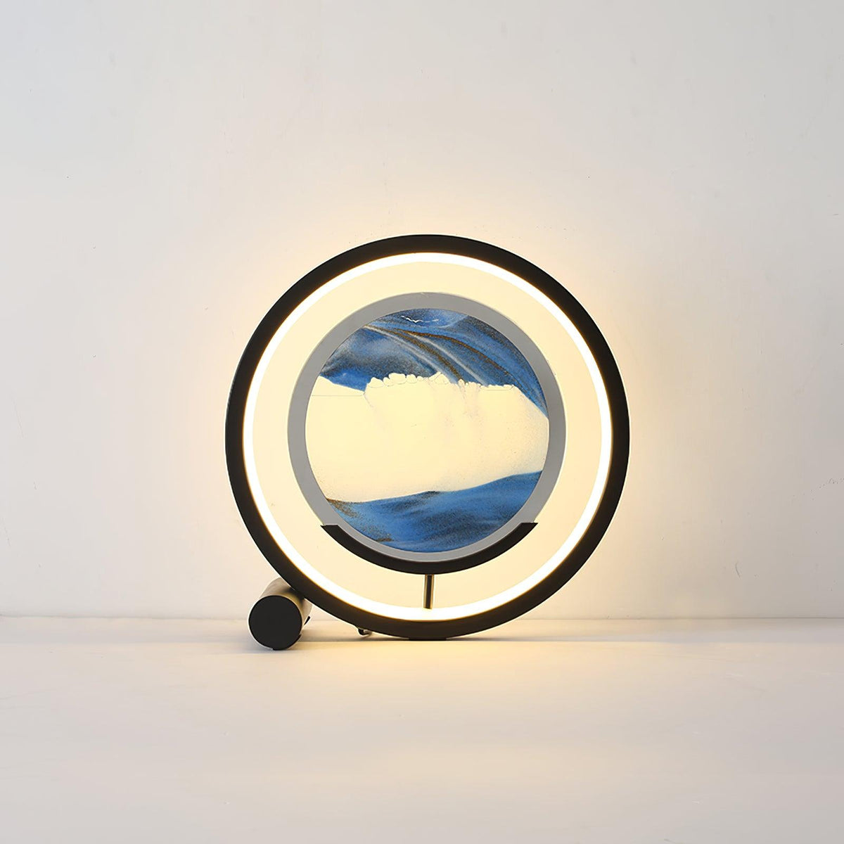 Sandrelle Table Lamp in black with lit LED ring around a round blue sand art panel on a plain backdrop