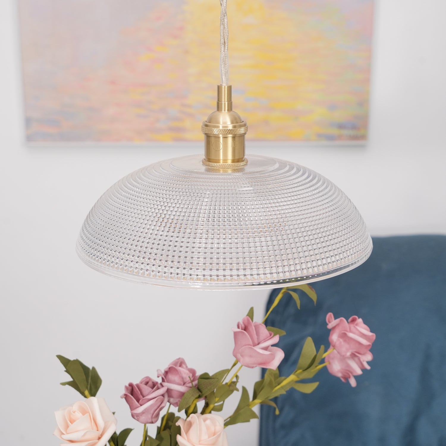Pendant light with a ribbed glass dome shade and a gold-toned lamp holder hanging above a vase of roses with a painting behind.