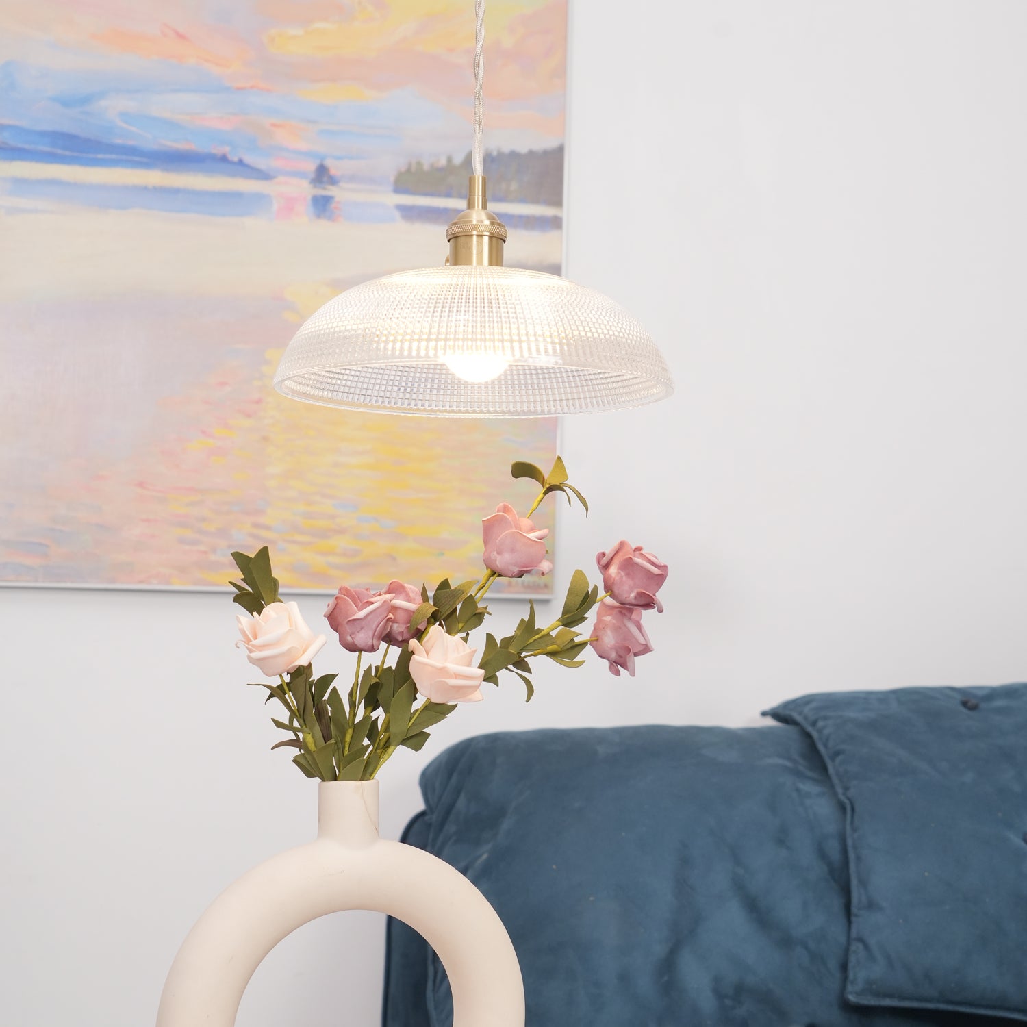 Pendant light with a ribbed glass dome shade and a gold-toned lamp holder, lit, above roses with a painting and a blue throw behind.