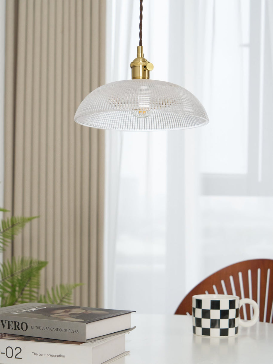 Pendant light with a ribbed glass dome shade and a gold-toned lamp holder, unlit, above books and a mug beside a ribbed curtain.