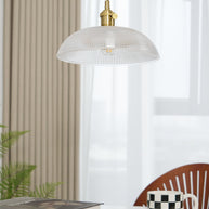 Pendant light with a ribbed glass dome shade and a gold-toned lamp holder, unlit, above books and a mug beside a ribbed curtain.