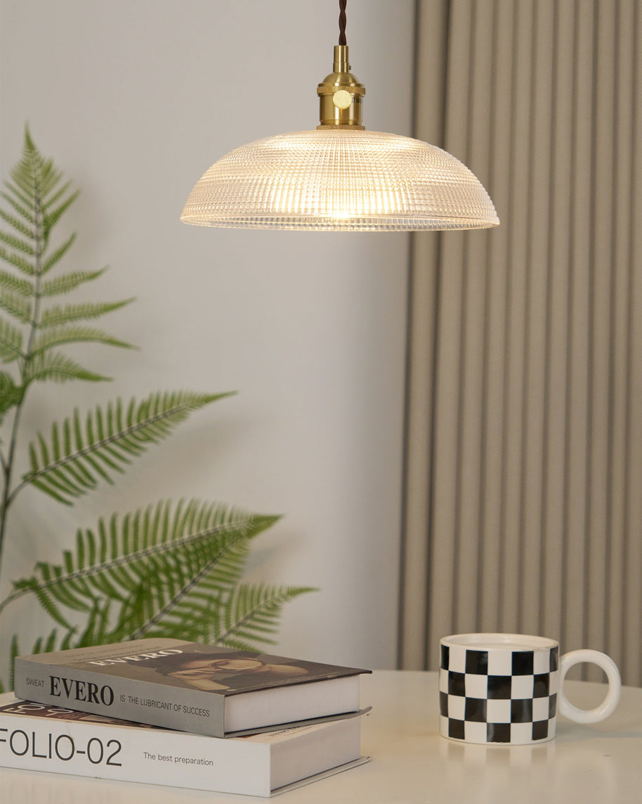 Pendant light with a ribbed glass dome shade and a gold-toned lamp holder, lit, above a table with a fern, books and a mug.