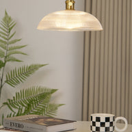 Pendant light with a ribbed glass dome shade and a gold-toned lamp holder, lit, above a table with a fern, books and a mug.