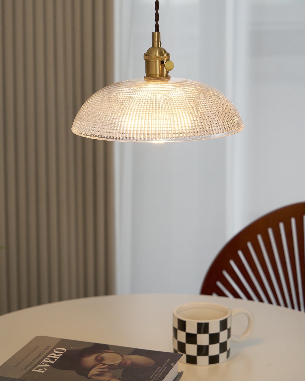 Pendant light with a ribbed glass dome shade and a gold-toned lamp holder, lit, above a table with a mug and a wooden chair behind.