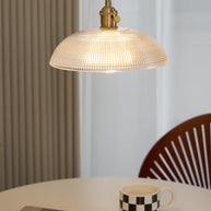 Pendant light with a ribbed glass dome shade and a gold-toned lamp holder, lit, above a table with a mug and a wooden chair behind.