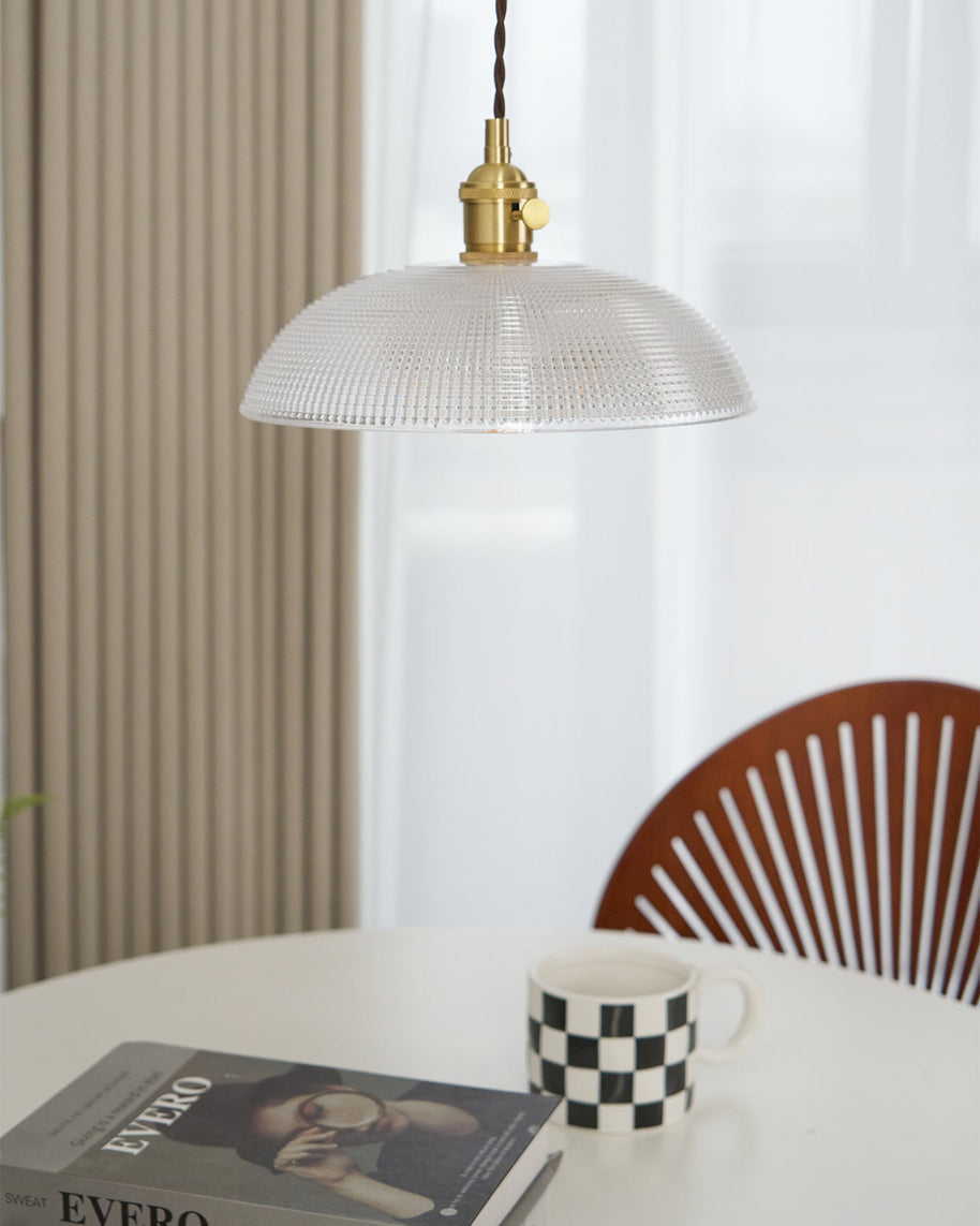 Pendant light with a ribbed glass dome shade and a gold-toned lamp holder, unlit, above a table with a mug and a wooden chair behind.