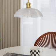 Pendant light with a ribbed glass dome shade and a gold-toned lamp holder, unlit, above a table with a mug and a wooden chair behind.