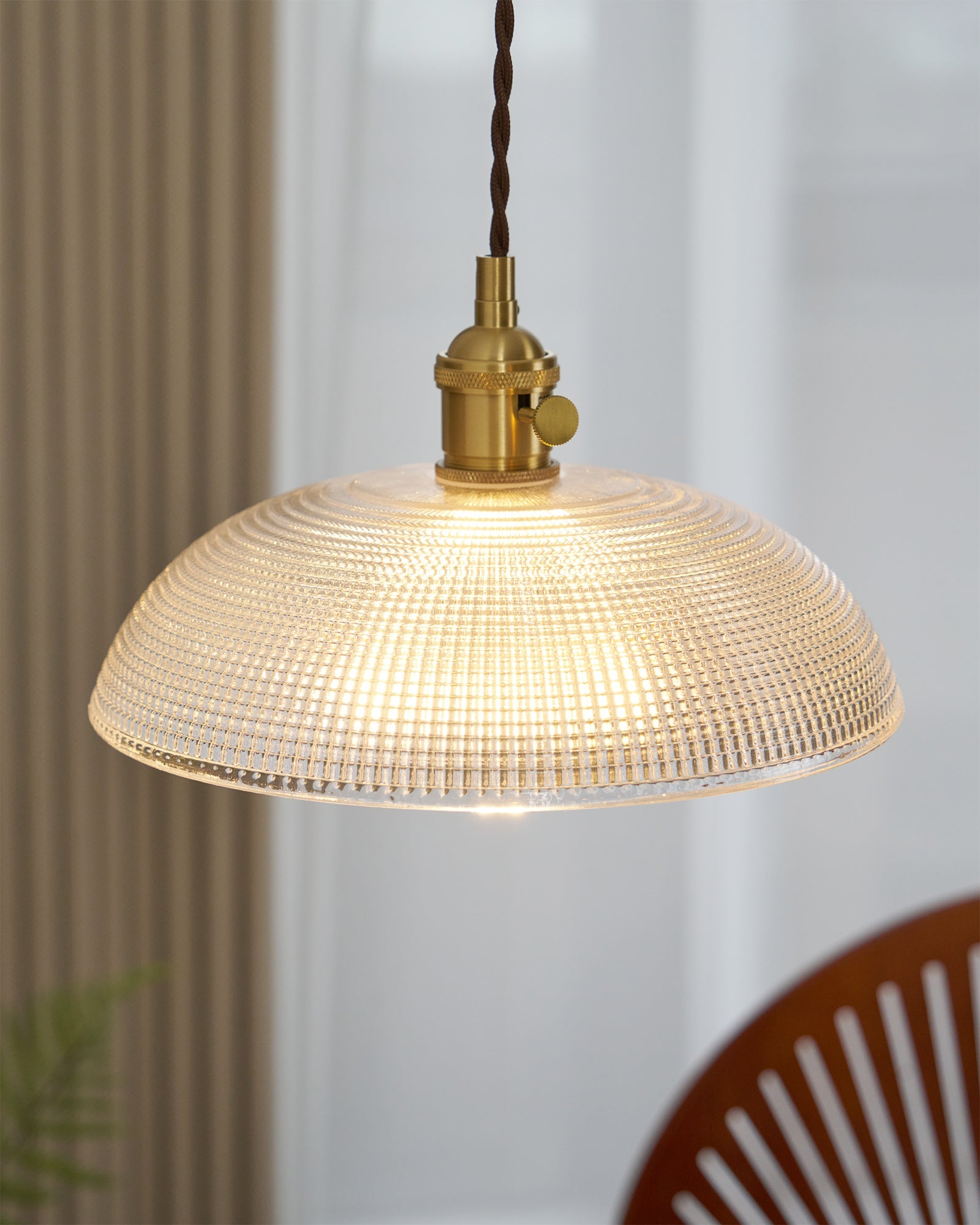 Pendant light with a ribbed glass dome shade and a gold-toned lamp holder, lit, above a table with a wooden chair behind.