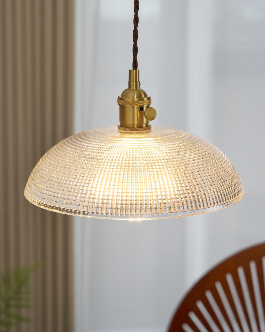 Pendant light with a ribbed glass dome shade and a gold-toned lamp holder, lit, above a table with a wooden chair behind.