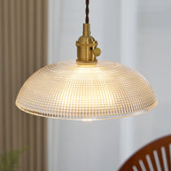 Pendant light with a ribbed glass dome shade and a gold-toned lamp holder, lit, above a table with a wooden chair behind.