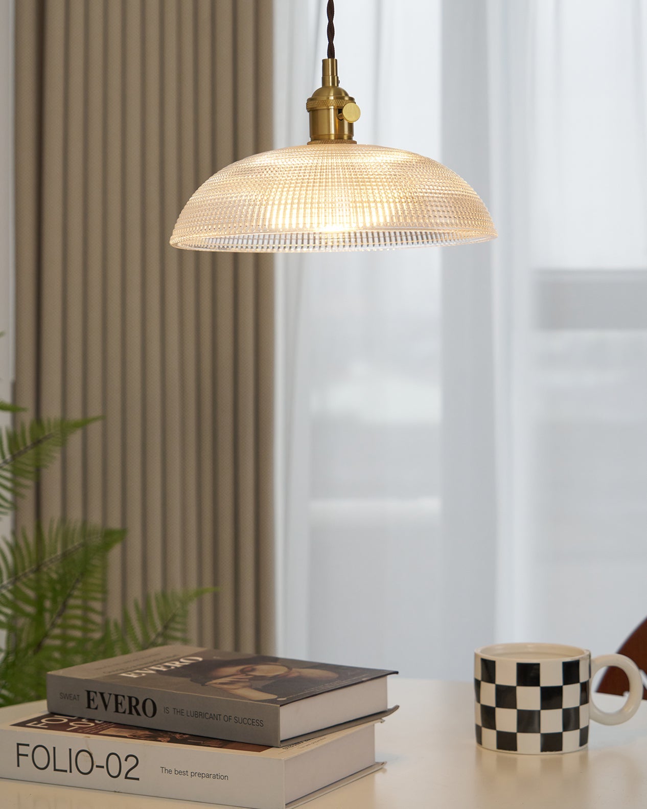 Pendant light with a ribbed glass dome shade and a gold-toned lamp holder, lit, above books and a mug beside a ribbed curtain.