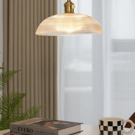 Pendant light with a ribbed glass dome shade and a gold-toned lamp holder, lit, above books and a mug beside a ribbed curtain.