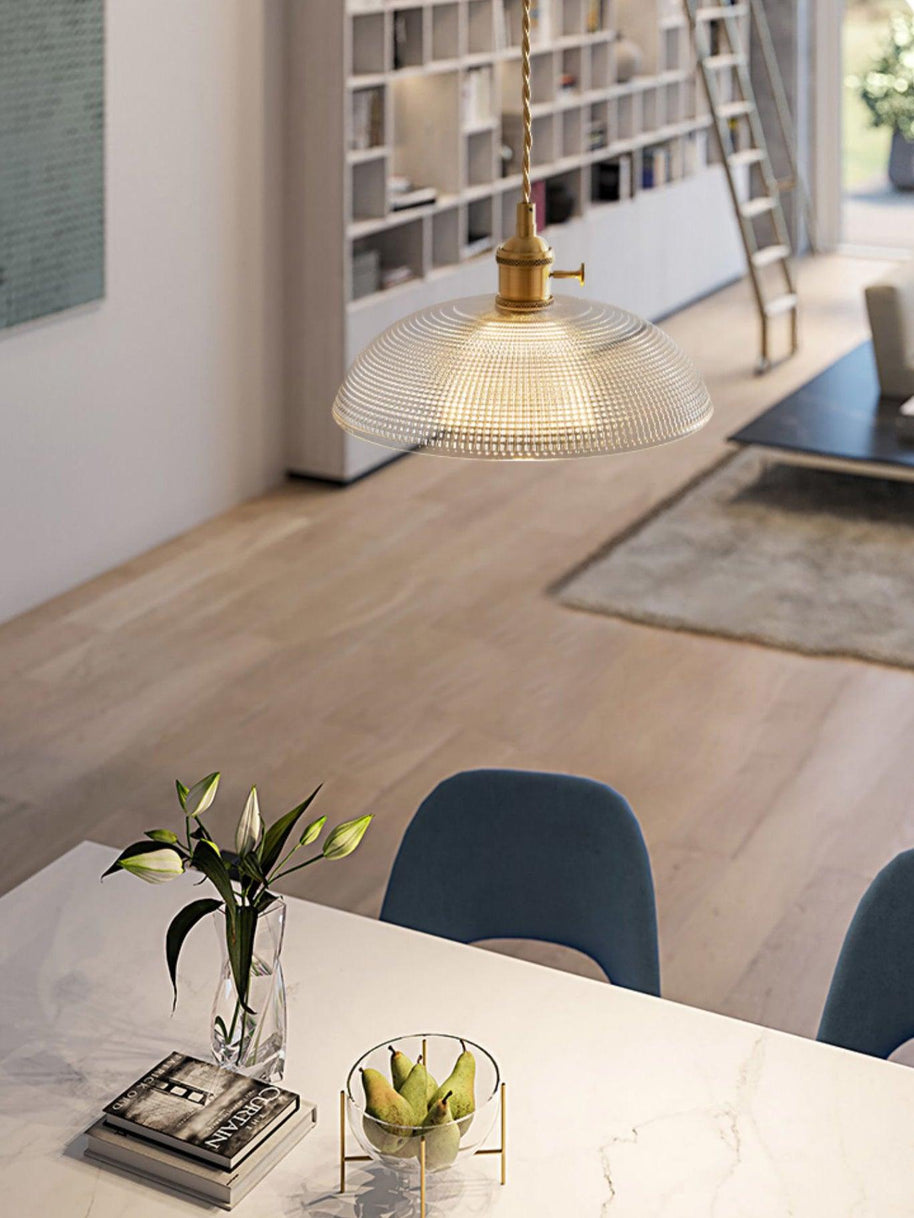 Pendant light with a ribbed glass dome shade and a gold-toned lamp holder, lit, hanging over a counter with books and a vase.