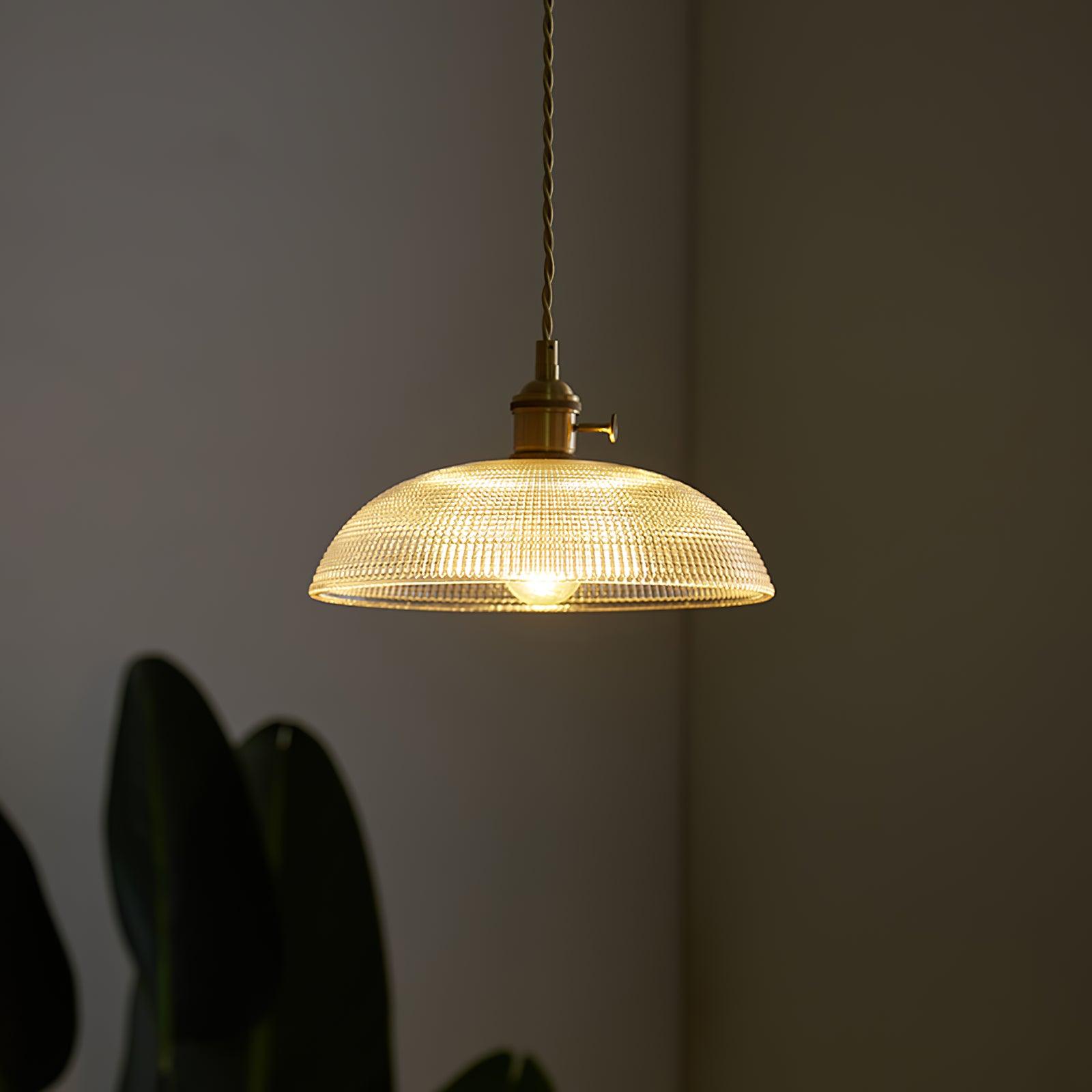 Pendant light with a ribbed glass dome shade and a gold-toned lamp holder, lit, hanging in a dim room with plants behind.