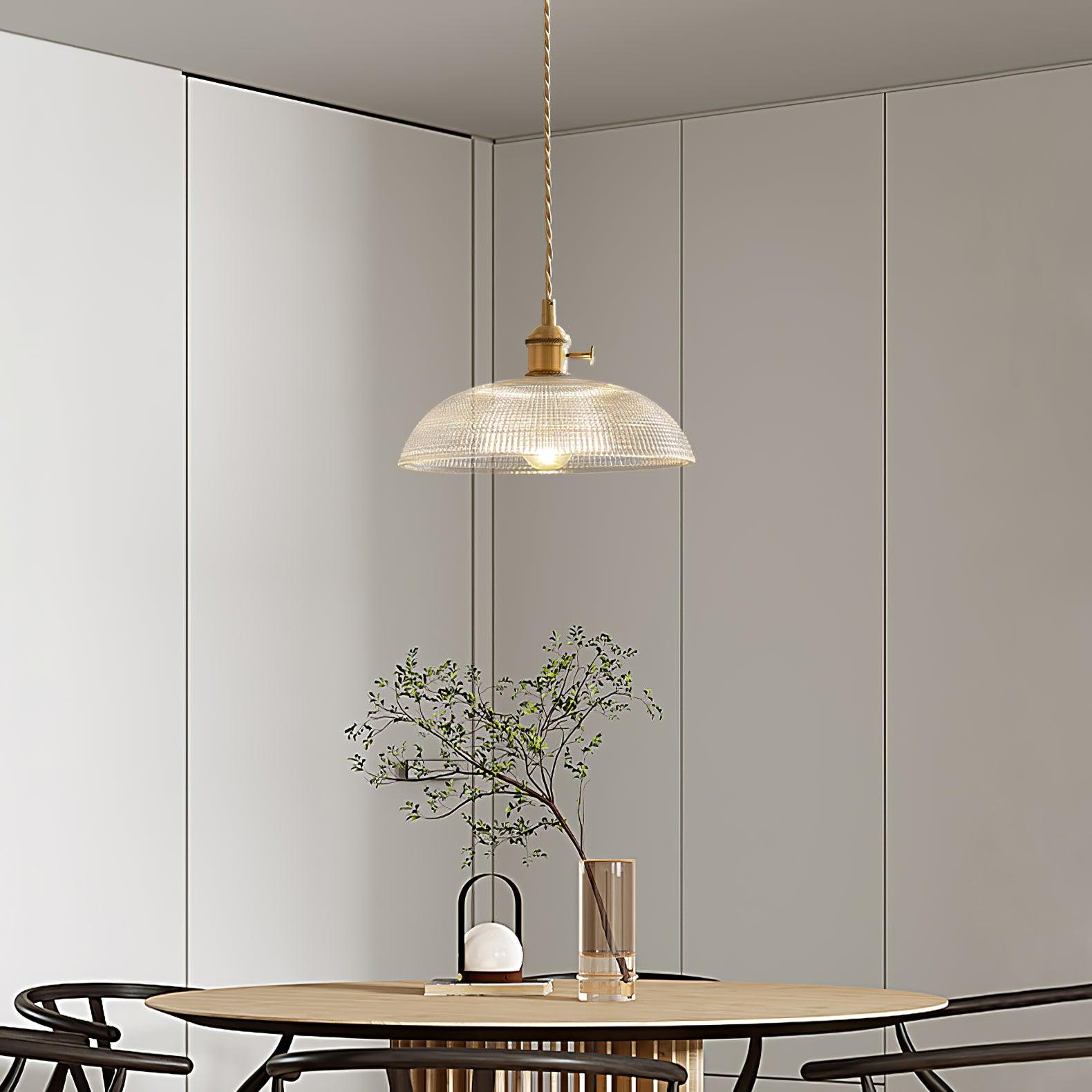 Pendant light with a ribbed glass dome shade and a gold-toned lamp holder hanging above a dining table in a pale room.
