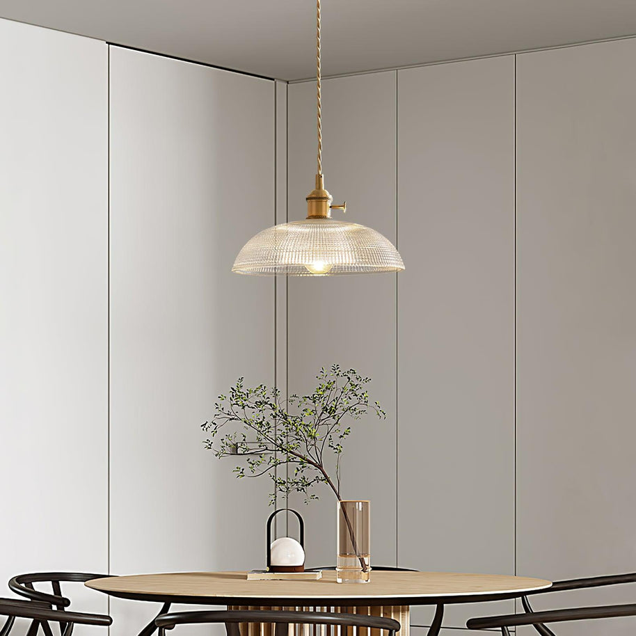 Pendant light with a ribbed glass dome shade and a gold-toned lamp holder hanging above a dining table in a pale room.