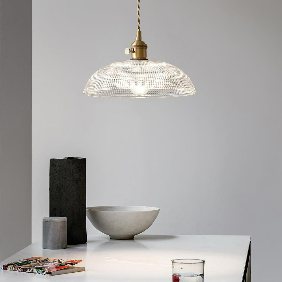 Pendant light with a ribbed glass dome shade and a gold-toned lamp holder hanging above a table set with a bowl and dark ornaments.